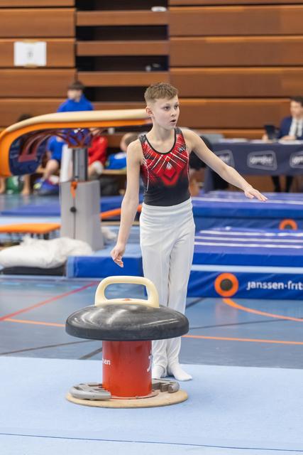 Young male gymnast in red and black leotard stands beside mushroom apparatus, arms extended, focused expression during training