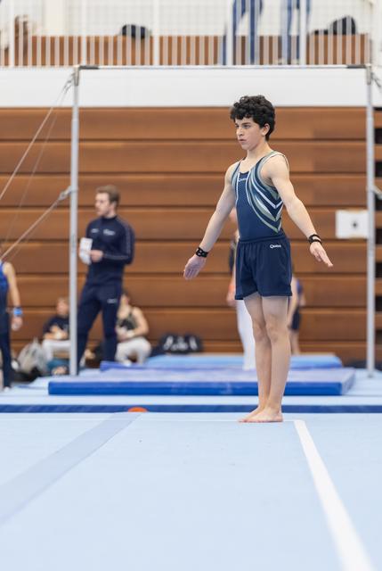 Young gymnast in navy leotard stands barefoot on floor mat with arms out, focused expression before routine
