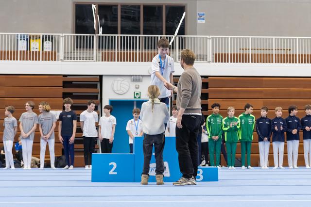 Coach presenting medal to young gymnast standing on first place podium as teammates watch from below in indoor sports hall