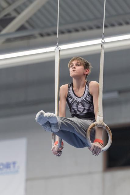 Young male gymnast performs L-sit on rings, demonstrating focus and strength in an indoor training facility