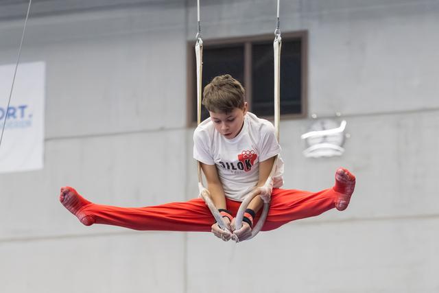 Young gymnast performs L-sit straddle hold on rings, displaying concentration and strength in training facility