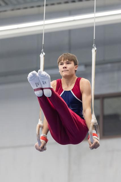 Young gymnast performing L-sit position on rings, displaying intense concentration and controlled form in gymnasium
