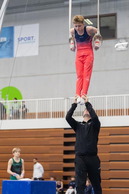 Young gymnast in coral gradient leotard performs on rings while coach supports from below in training gymnasium