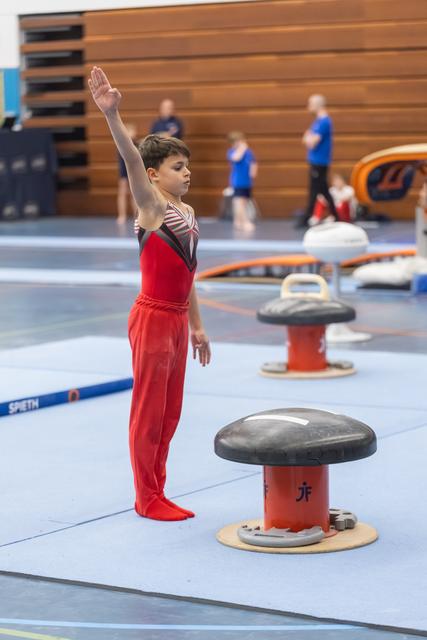 Young gymnast in red uniform stands in salute position beside pommel horse, displaying perfect form during training session