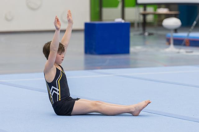Young gymnast performs a seated flexibility stretch with arms raised overhead on the floor exercise mat in training facility
