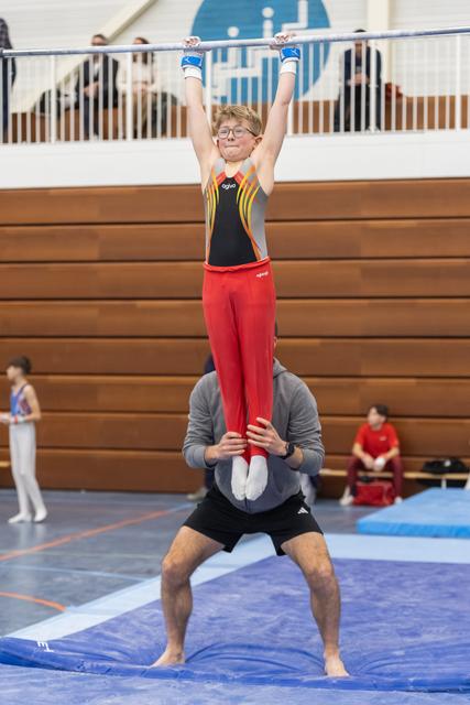 Young gymnast in red pants performs shoulder stand on adult spotter wearing grey, gripping bar overhead in gymnasium