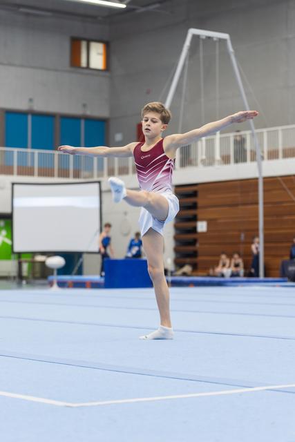 Young gymnast performing a split leap during floor routine at indoor gym, arms extended with focused expression