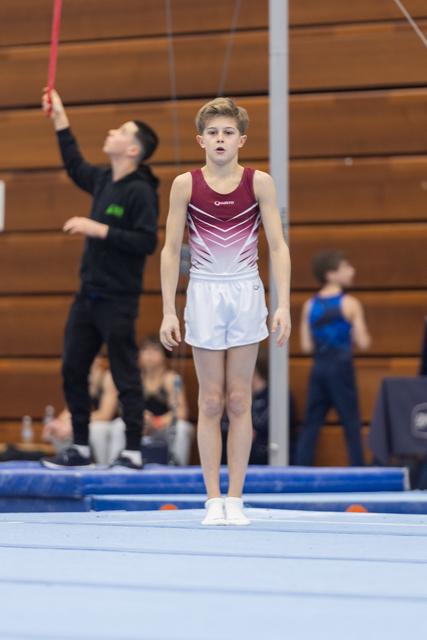 Young gymnast in burgundy and white leotard stands focused on the floor mat, coach visible in background