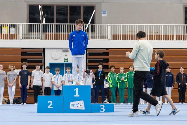 Young gymnast in blue stands atop first place podium while coach and another child walk past during medal ceremony