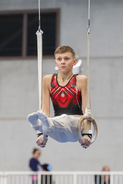 Young gymnast performs on still rings with intense focus, wearing red and black leotard in training facility
