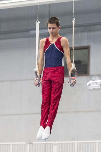 Young gymnast performing on still rings, focused expression, wearing red and navy uniform in training facility