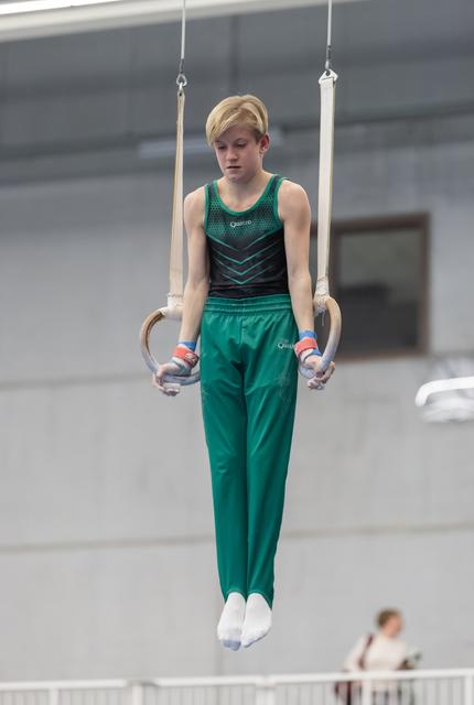 Young gymnast with blonde hair holds steady on still rings, focused expression, wearing green competition uniform in training hall