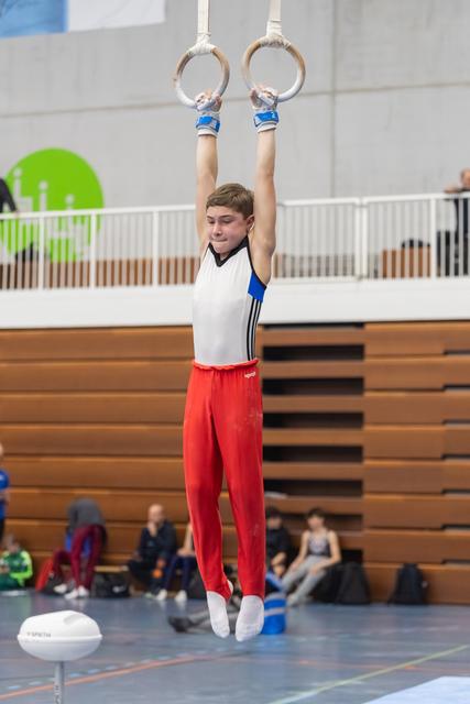 Young gymnast performing a still rings routine, arms extended overhead, wearing red pants and white top in an indoor gym