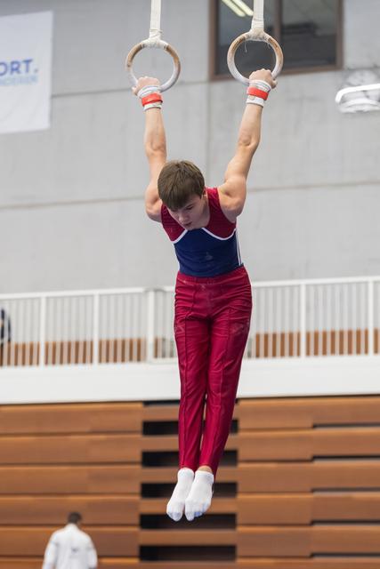 Young gymnast performs a still rings hold, arms extended overhead, wearing maroon and blue uniform in indoor gymnasium