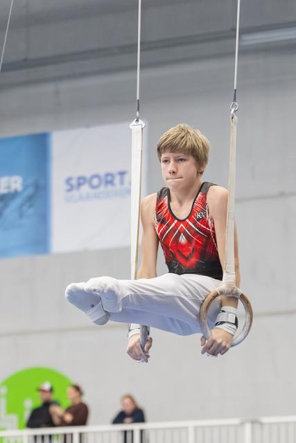 Young gymnast performing L-sit position on rings, displaying focused concentration in red and black leotard at indoor facility