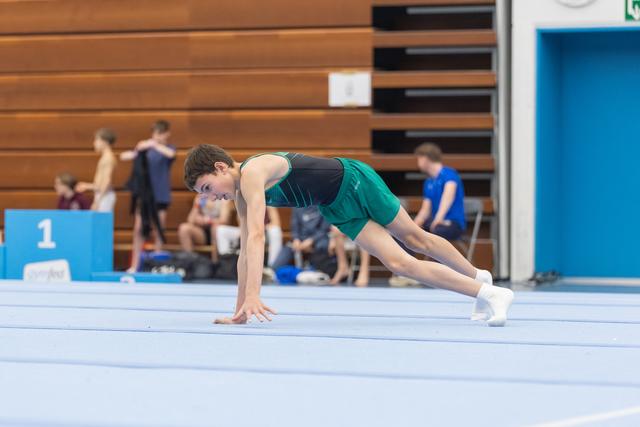 Young gymnast in teal and green uniform performs a deep lunge stretch on floor mat during warmup, focused and preparing