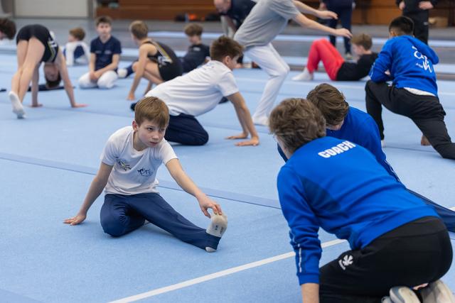 Young gymnast in white shirt stretches on blue mat while coach in blue jacket provides hands-on guidance and support