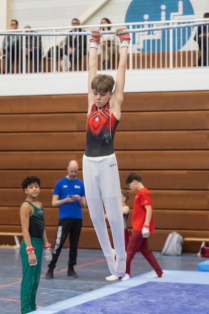 Young gymnast in red and black leotard performs vertical jump with arms raised high, teammates visible in background