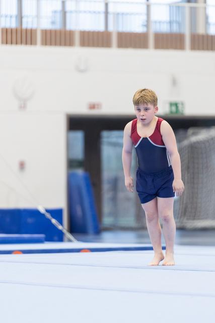 Young gymnast in navy and red leotard walks barefoot on floor mat, focused expression, training facility with blue equipment