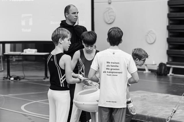 Young gymnasts gather around chalk bowl while coach observes, one wearing Team Josse shirt in gymnasium