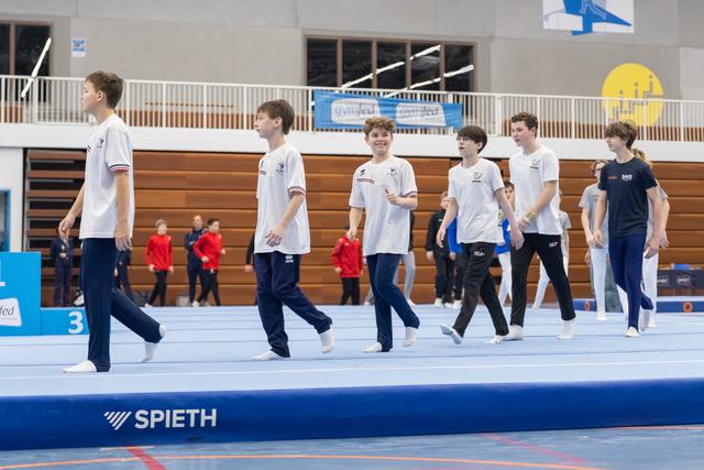 Six young male gymnasts in white team uniforms march in formation across a blue Spieth floor mat during a team presentation