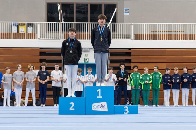 Young gymnasts stand on award podium with medals while teammates line up behind them in gymnasium