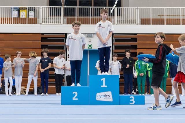 Two young gymnasts stand on first and second place podium positions while others gather around in a gymnasium