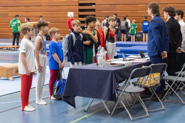 Group of young male gymnasts in sportswear line up at a registration table, engaging with officials in an indoor facility