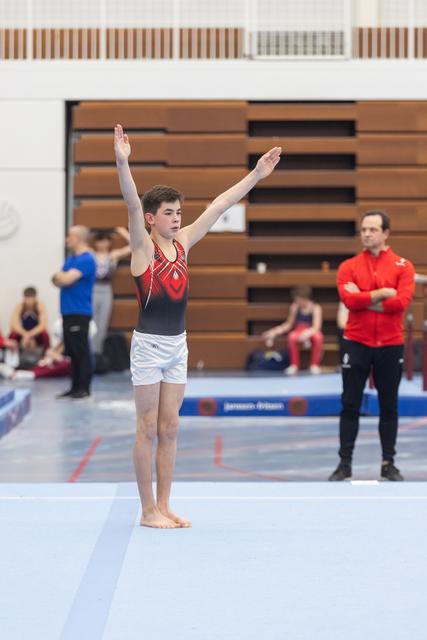Young gymnast in red and black leotard stands with arms raised, preparing for floor routine in gymnasium