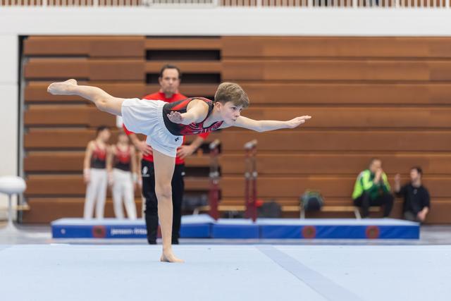 Young gymnast performs an arabesque balance on floor exercise, extending his leg high while his coach observes from behind