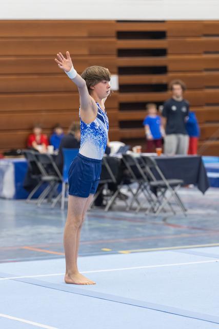 Male gymnast in blue and white leotard stands with arm raised in starting position on floor mat at indoor meet