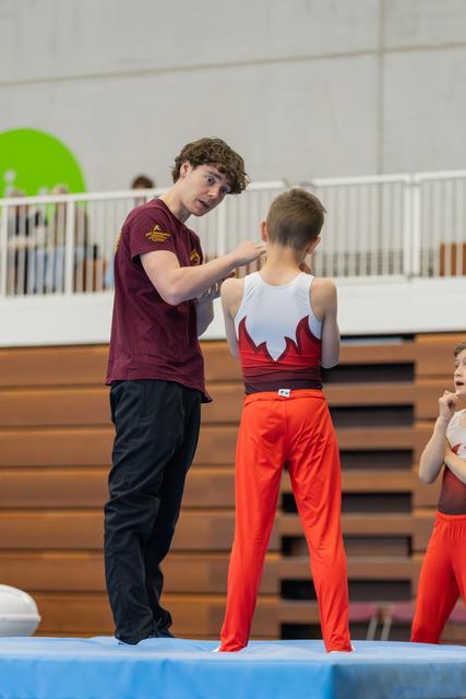 A teenage coach attentively gives feedback to a young male gymnast in a red and white leotard at an indoor gymnastics event.