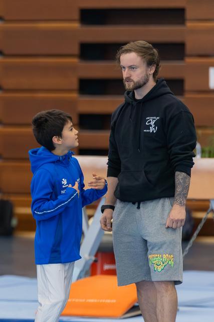 A young gymnast in a blue hoodie speaks earnestly to his coach, who listens attentively in a gymnasium setting.