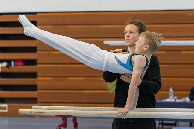 A teen coach spots a young boy performing a horizontal hold on parallel bars, both focused and composed in a gymnasium.