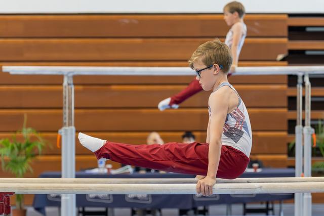 Focused young gymnast with glasses performs a straddle hold on parallel bars, legs extended forward, intense concentration.