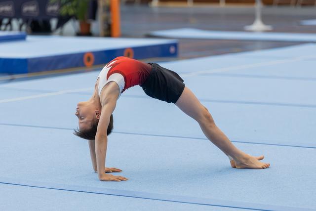 Young gymnast performs a bridge pose on blue floor mat, head inverted, body arched with focused control.