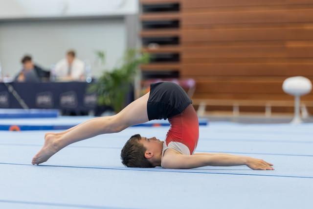 Male gymnast performs a dramatic backbend arch on the floor mat, legs extended overhead, in a competitive gymnastics hall.