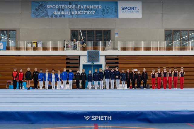 Young male gymnasts in team uniforms stand in a long lineup on the floor exercise mat at a Sport Vlaanderen event.