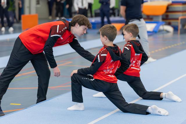 A teenage coach attentively guides two young boys doing splits stretches on a blue gymnastics mat, all in red uniforms.