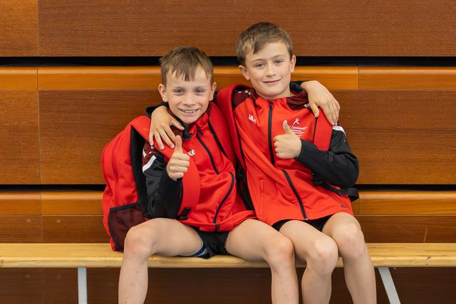 Two young boys in red sports jackets sit on a bench, arms around each other, both giving enthusiastic thumbs up and smiling.