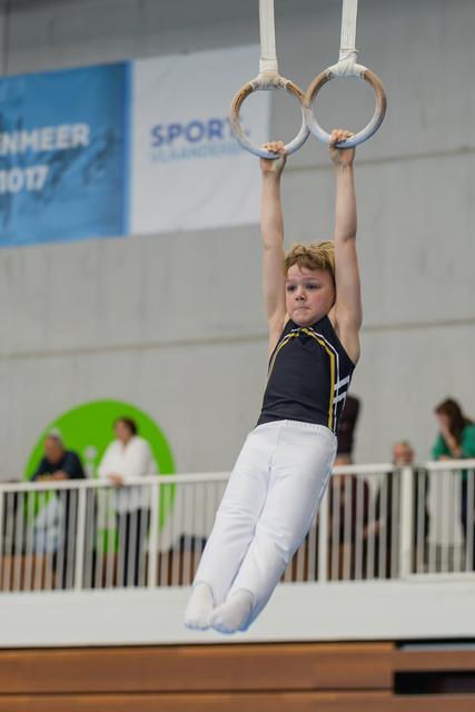 Determined young gymnast hangs from still rings, legs together, intense focus on his face during an indoor competition.