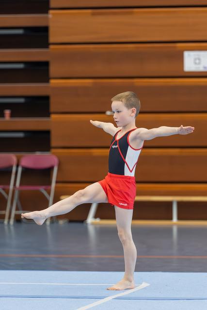 Young boy gymnast balances on one leg with arms outstretched, focused and composed during a floor routine in a sports hall.