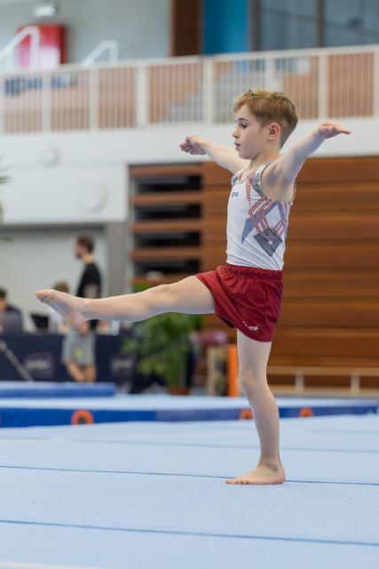 Young male gymnast performs an arabesque on the floor exercise mat, arms extended with focused expression.