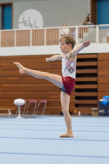Focused young male gymnast performs a leg raise on the floor exercise, arms outstretched for balance in an indoor gym.