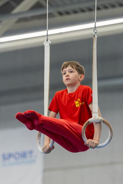 Young boy in red grips gymnastics rings mid-routine, legs raised, expression calm and concentrated in a sports hall.