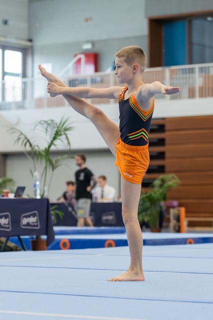 Young boy gymnast performs a precise standing leg raise on the floor mat, arms extended, focused expression.