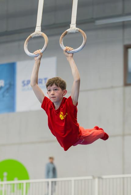 Focused young boy in red kit hangs from gymnastic rings mid-routine at an indoor sports hall competition.