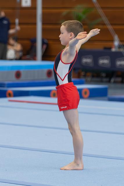 Young male gymnast stands tall on the floor mat, arm raised in a proud opening salute during a gymnastics meet.