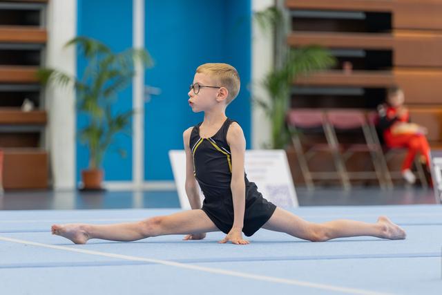 Young boy in black leotard performs perfect splits on blue floor mat, gazing ahead with concentration and glasses.