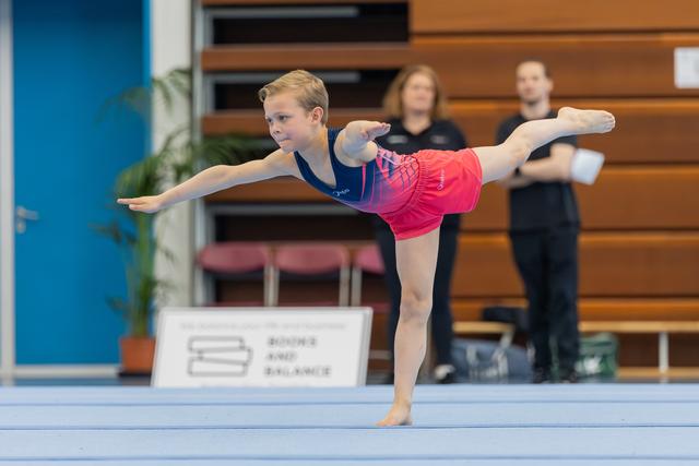 Young gymnast performs a precise arabesque on the floor, arms outstretched, focused expression, judges watching in background.